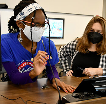 A Classroom support student assists an instructor, demonstrating how to set up the built in microphone for use. 