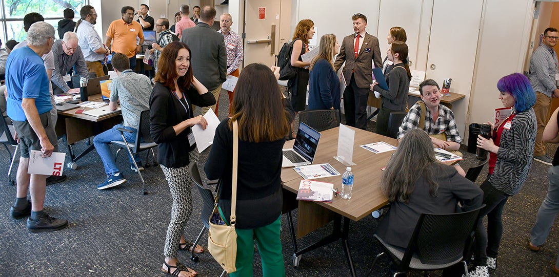 A large room is full of people gathered around tables with laptops and handouts, talking animatedly in small groups.