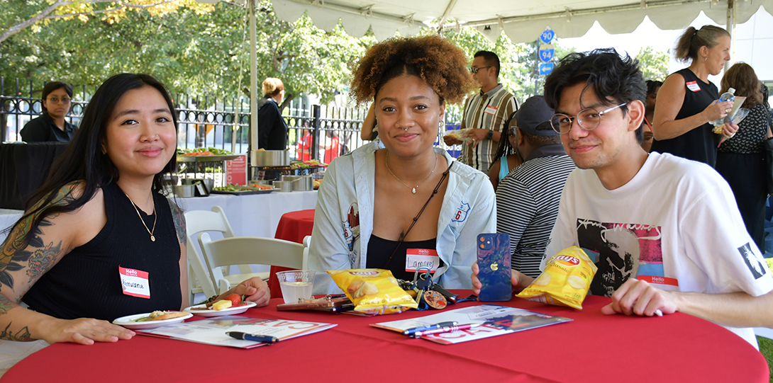 three young adults sit at a round table snacking on chips and fruit, smiling for the camera. Other people in the background help themselves from a buffet and mingle.